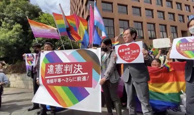 名古屋地裁前で「違憲判決　婚姻平等へさらに前進！」と書いた旗を掲げる弁護団。弁護団の後ろには、さまざまなフラッグを掲げる支援者や他地域の弁護団・原告が並んでいる