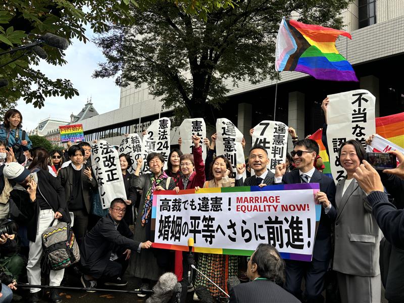 東京高裁の正門前にて「高裁でも違憲！婚姻の平等へさらに前進！」と書いた横断幕を掲げる「結婚の自由をすべての人に」東京１次原告(控訴人)と弁護団や支援者達と、それを取り囲む大勢の報道陣の様子の写真