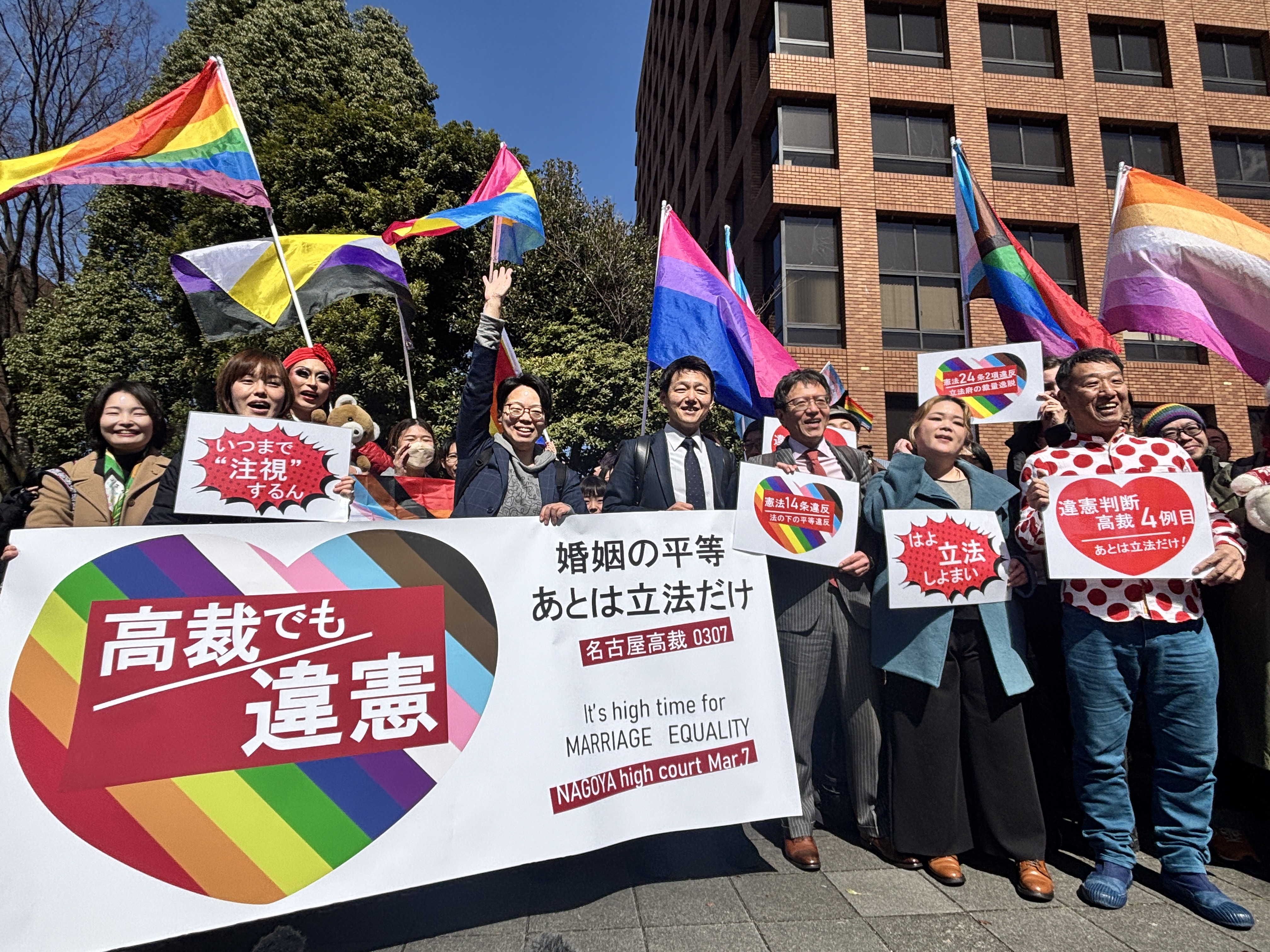 名古屋高裁前での旗出しの様子。 愛知弁護団が旗を掲げており、旗には「高裁でも違憲、婚姻の平等あとは立法だけ　名古屋高裁０３０７」と書かれている。 弁護団の周りには、他地域の原告や弁護団、支援者たちがいる。