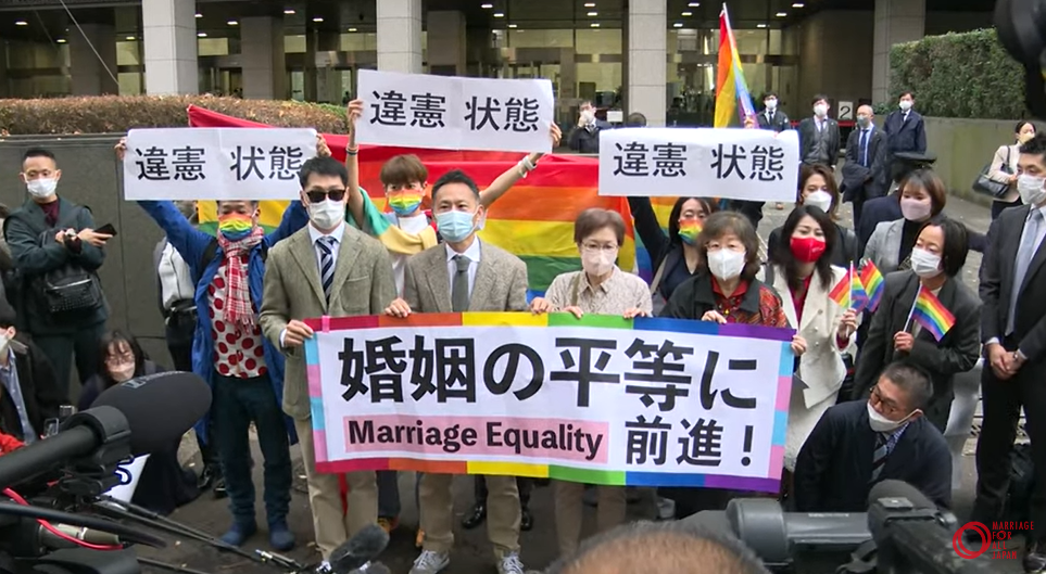 ①	判決言渡し後、裁判所前で、たくさんの報道陣と支援者に対し、原告らが「婚姻の平等に前進」と書かれた大きな旗と「違憲状態」と書かれた紙を掲げました。