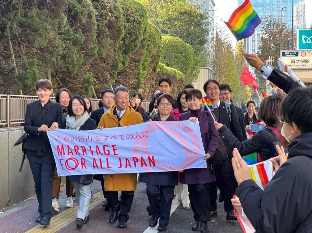 東京地方裁判所前で旗を持ちながら行進する原告らと弁護団員