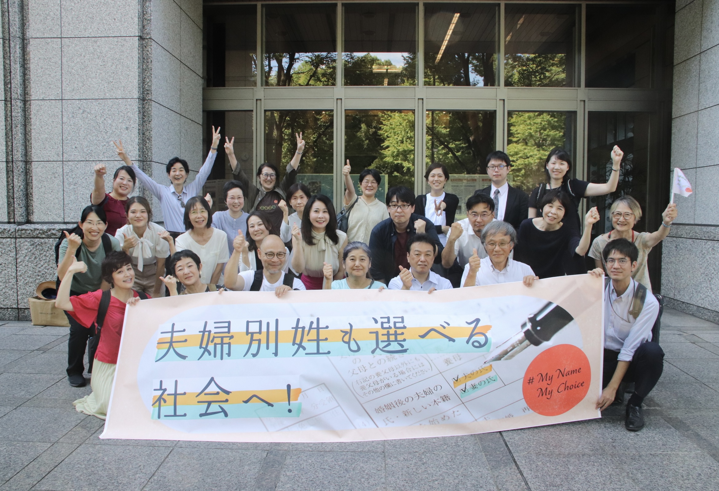 期日前の原告・弁護団らの集合写真。弁護士会館前にて。夫婦別姓も選べる社会へ、の横断幕を掲げる。
