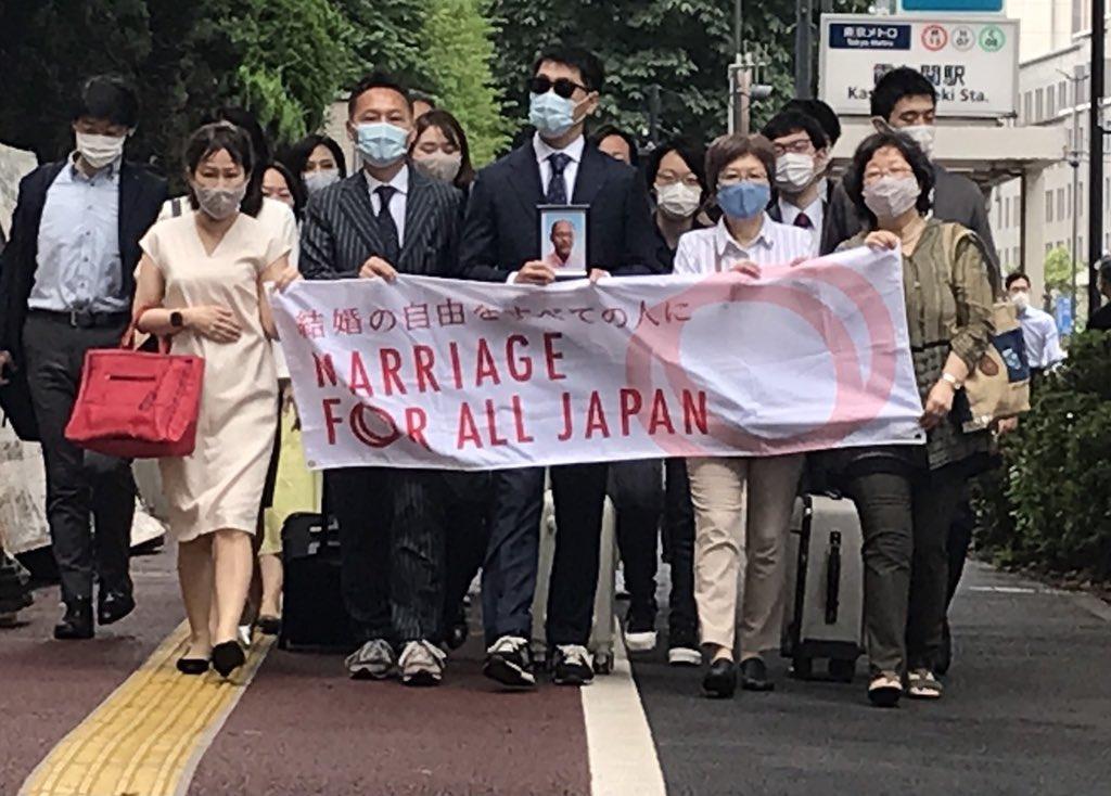 歩いていてこちらに向かってくる大勢の人たち。1列目は５人の人がMarriage For All Japan – 結婚の自由をすべての人にと書かれた横断幕を持っている。真ん中の男性が、小さな写真（佐藤郁夫さんが写っている）を持っている。