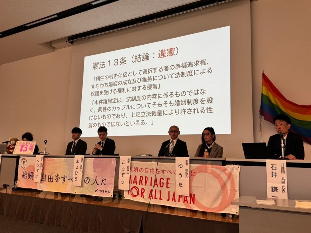 報告会の様子。原告さん4名と弁護団共同代表が壇上に並んでいる。投影スライドには、次のことが書かれている。 憲法13条　結論違憲 「同性の者を伴侶として選択する者の幸福追求権、すなわち婚姻の成立及び維持について法制度による保護を受ける権利に対する侵害」 「本件諸規定は、法制度の内容に係るものではなく、同性のカップルについてそもそも婚姻制度を設けないものであり、上記立法裁量により許される性質のものではないといえる。」 
